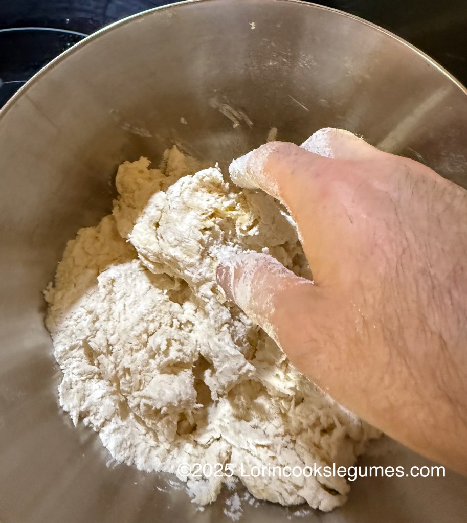 A hand mixing dough in a metallic bowl, showing a textured combination of flour and ingredients for making plantain dumplings.