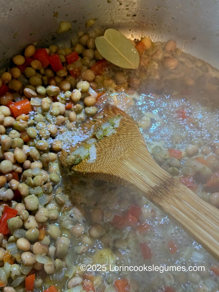 Close-up of a pot containing cooked pigeon peas, diced red bell peppers, and a bay leaf, with a wooden spoon stirring the mixture.