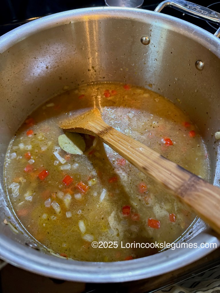 A pot of asopao soup cooking on the stove, featuring diced red bell peppers, onions, and a bay leaf, with a wooden spoon stirring the ingredients.