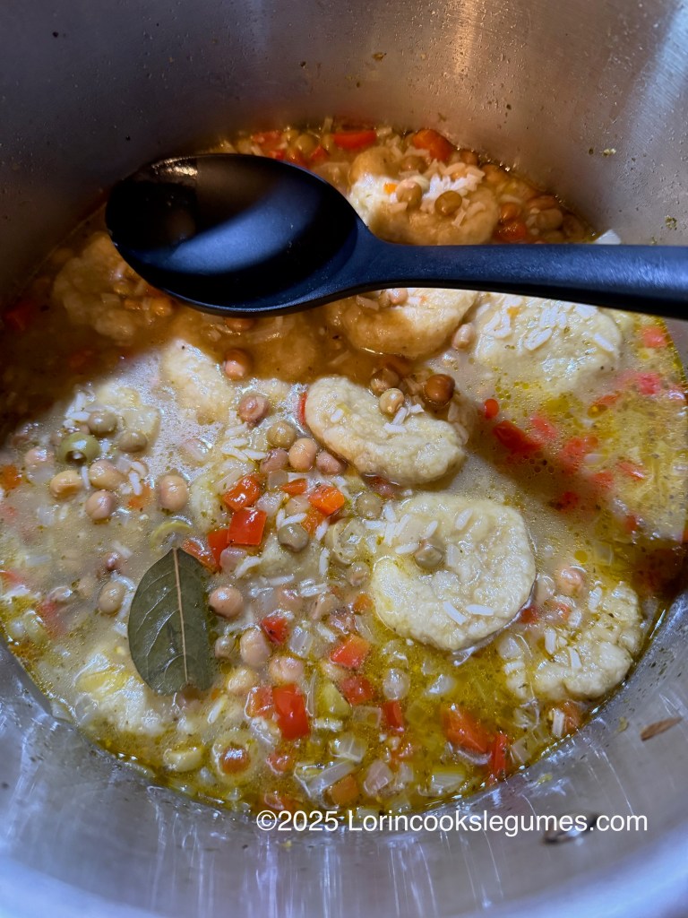 A pot of asopao soup with dumplings, featuring pigeon peas, diced red bell pepper, bay leaf, and spices. A black spoon rests on the edge of the pot.