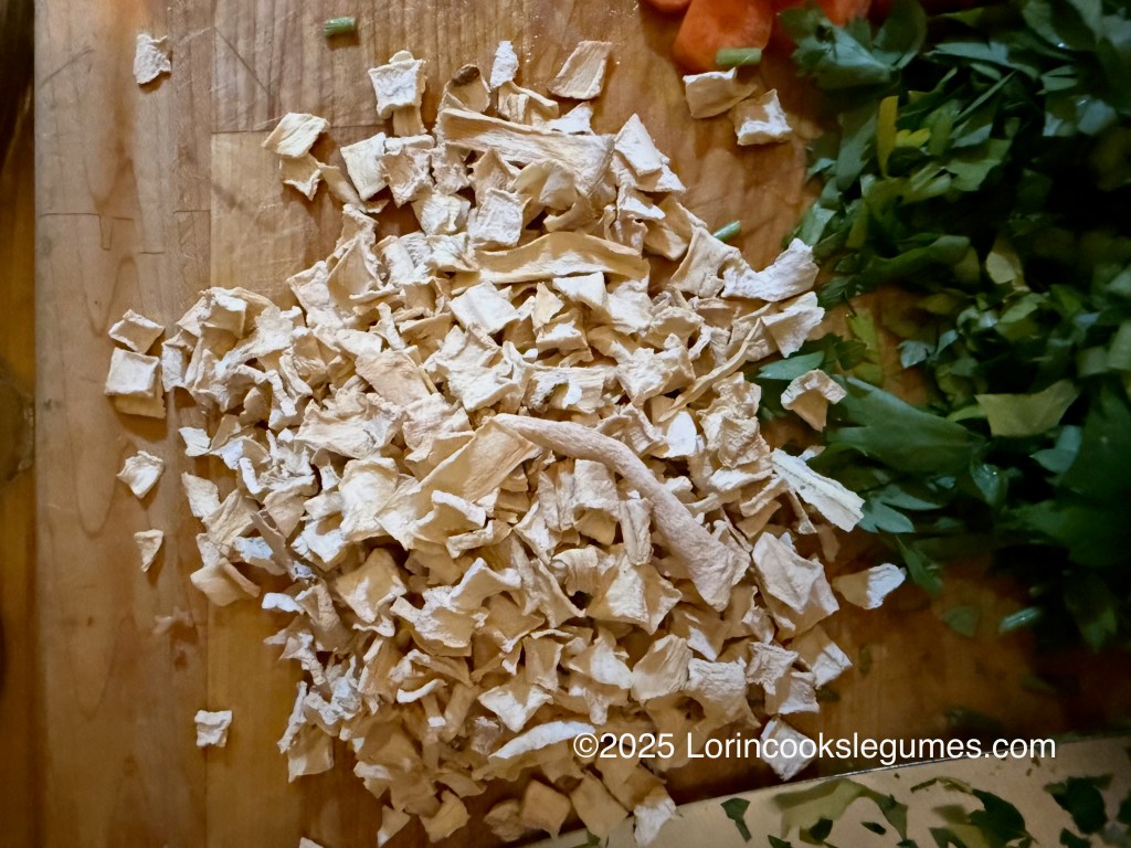 A pile of dried parsley root pieces on a wooden cutting board, with chopped fresh parsley and diced carrots visible beside it.