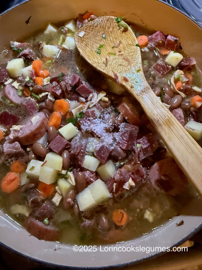 A pot of Fazulnica soup, filled with smoked meats, beans, potatoes, and colorful vegetables, stirred with a wooden spoon. A sprinkling of salt is visible on the surface. 