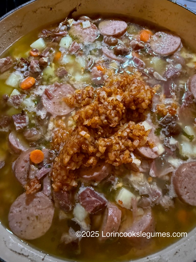 A pot of Fazulnica soup filled with diced smoked meats, vegetables, and a rich broth, with the onion-flour-paprika roux mixture on top, ready to be mixed in. 