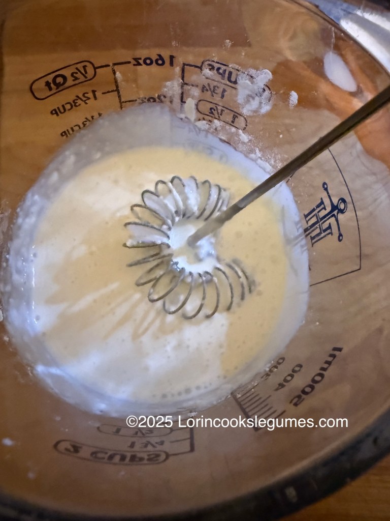 A glass measuring cup containing a mixture of cream and flour, being whisked with a wire whisk.