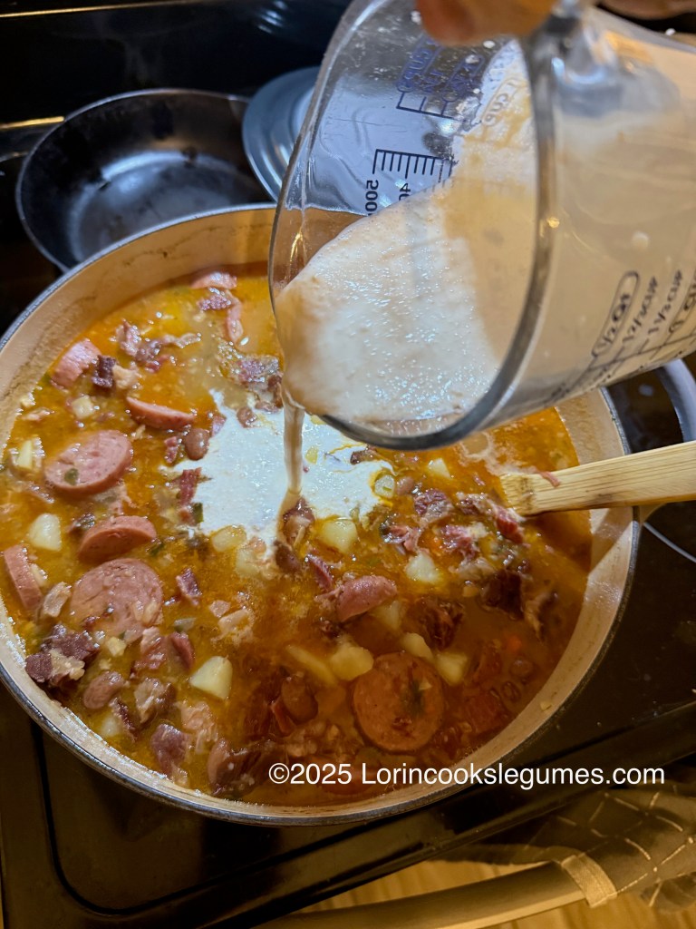 A hand pouring cream from a measuring cup into a pot of simmering Fazulovica soup, featuring smoked meats, beans, and vegetables.