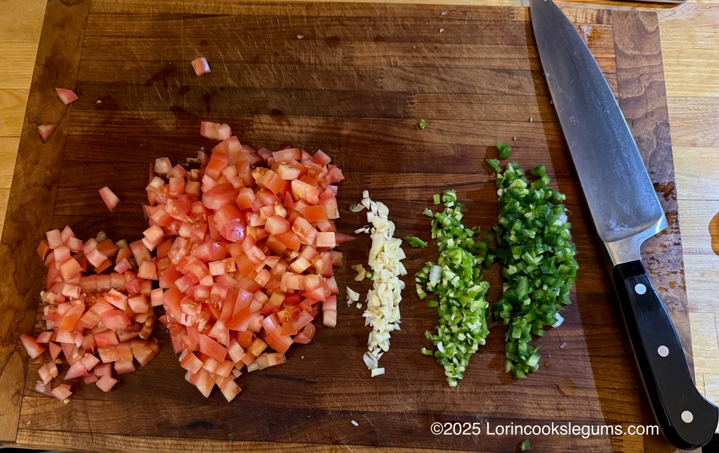 Chopped tomatoes, garlic, and green chili peppers arranged on a wooden cutting board, with a knife beside them.