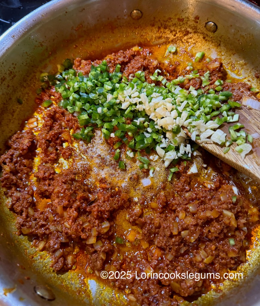 A pan with cooked chorizo, diced green chili peppers, and minced garlic is shown. The ingredients are being mixed together with a wooden spoon.