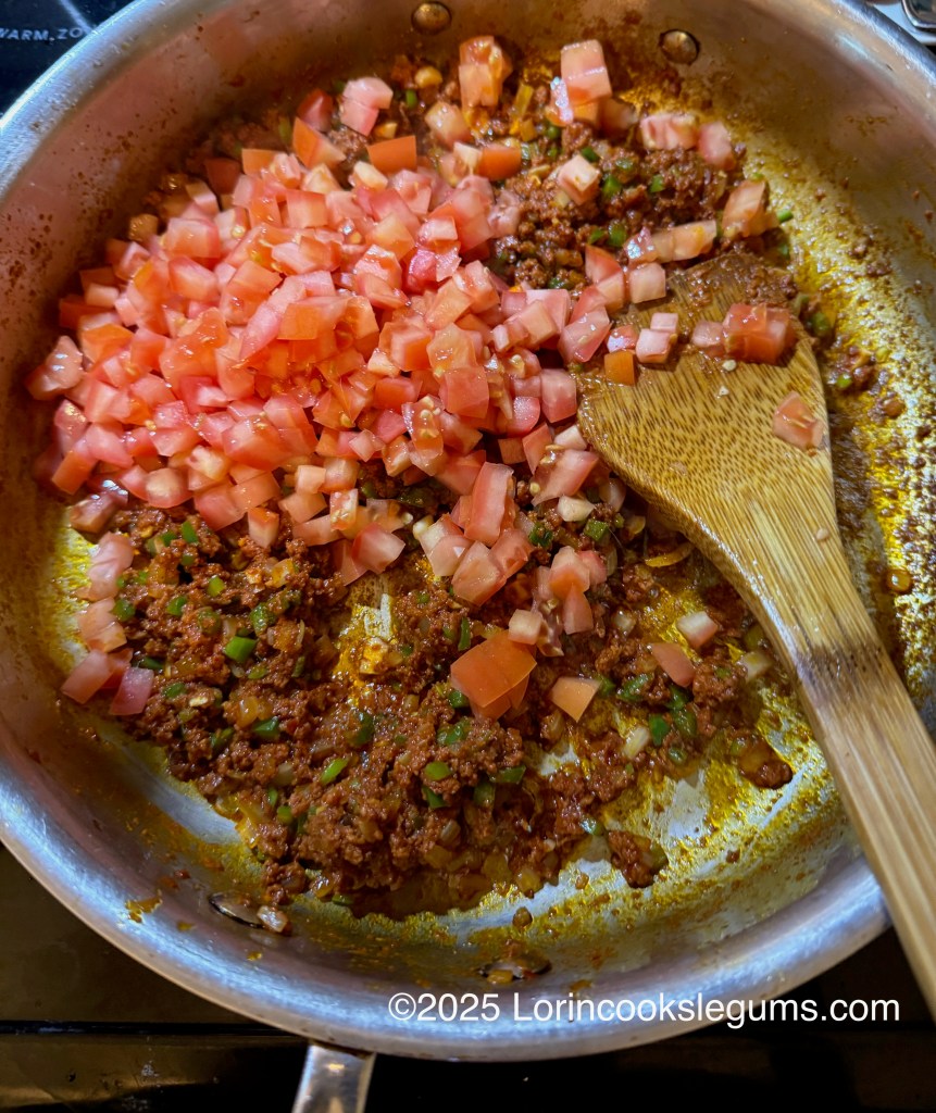 A frying pan containing sautéed chorizo with diced tomatoes and green chili peppers, stirred with a wooden spoon.