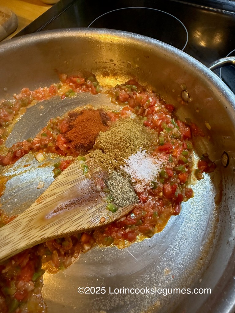 A close-up view of a skillet containing chopped tomatoes and pepper, with spices  about to be stirred in with a wooden spoon, showcasing a mixture of colors and textures.