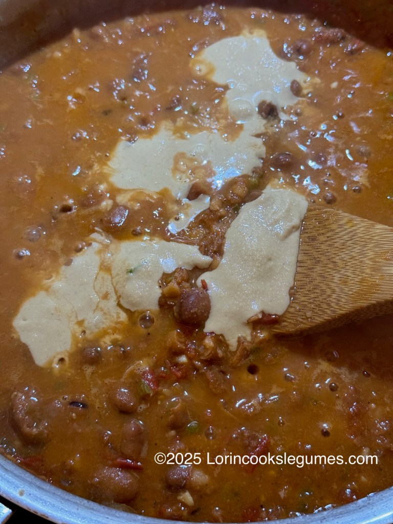 A close-up view of a pot of ful medames, a stew made from fava beans, showing a thick, creamy consistency with tahini being stirred in.
