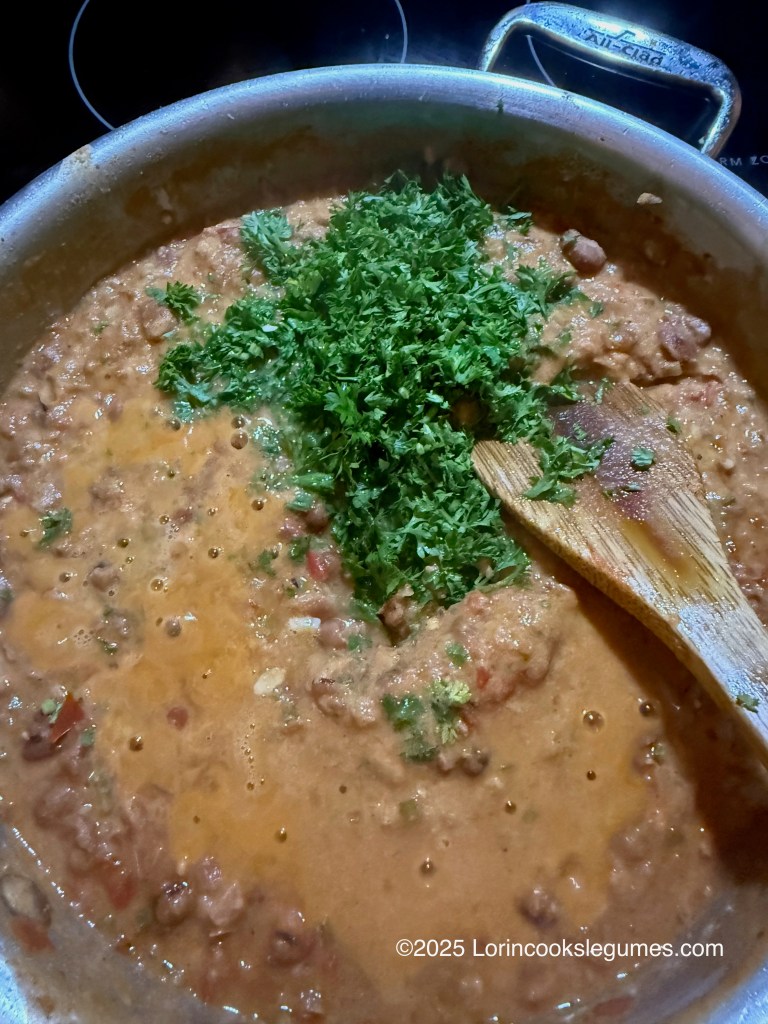 A pot of ful medames stew with a large handful of chopped parsley on top, stirred with a wooden spoon.
