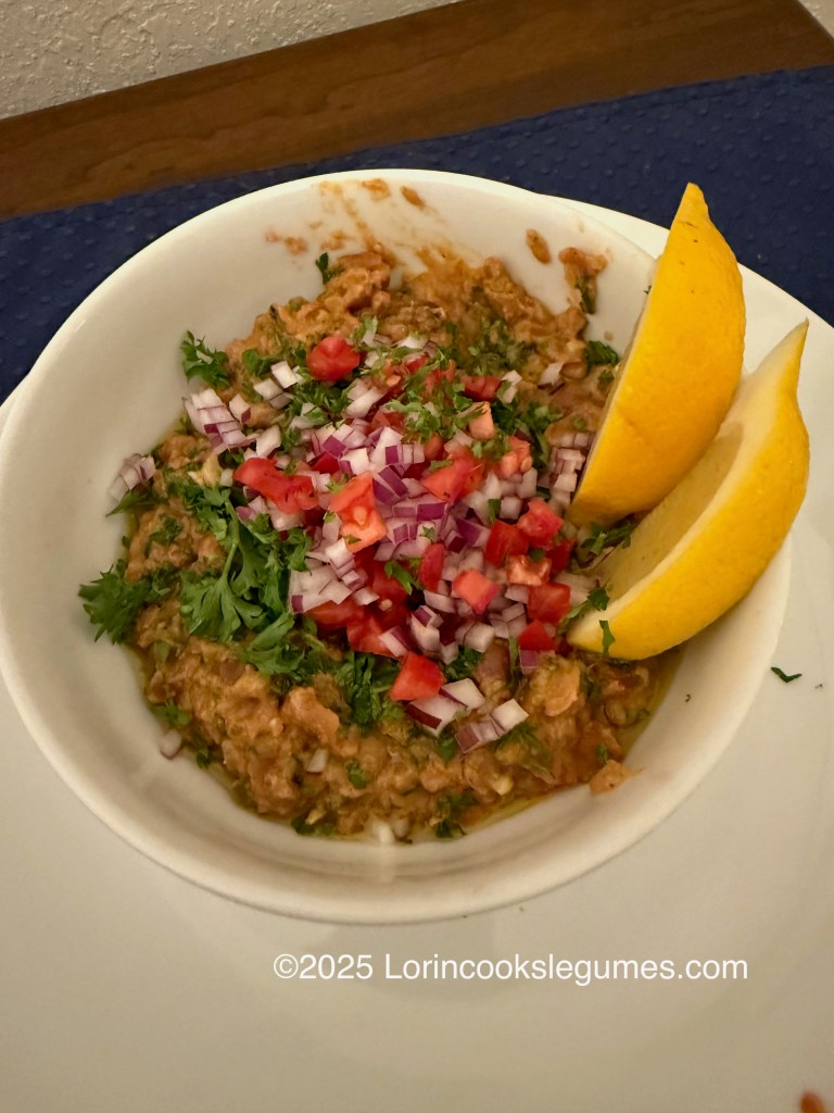 A bowl of ful medames topped with chopped tomatoes, onions, and parsley, accompanied by two lemon wedges.