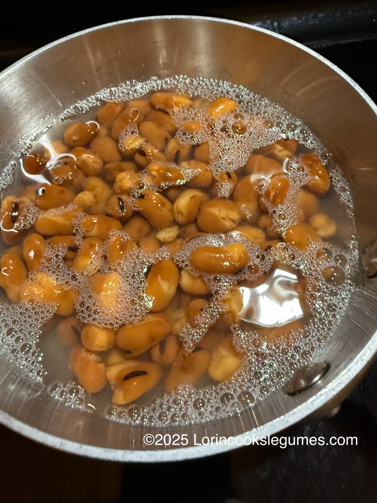 A pot filled with simmering fava beans in water, showing bubbles on the surface.