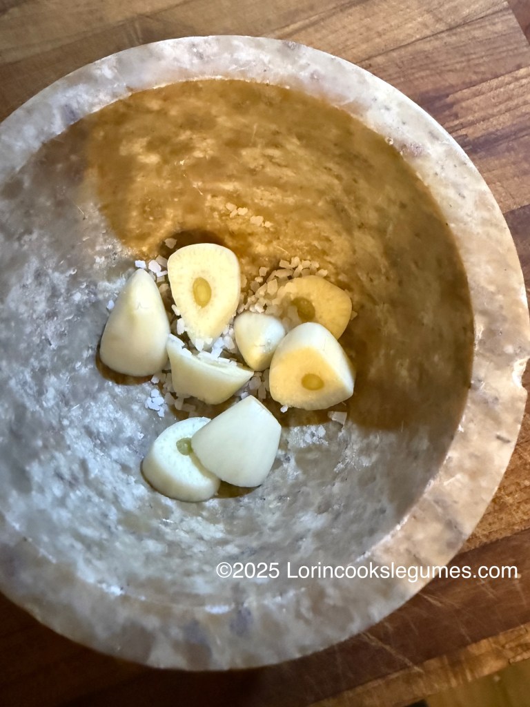 A stone mortar with several garlic cloves and a pinch of salt inside, resting on a wooden surface.