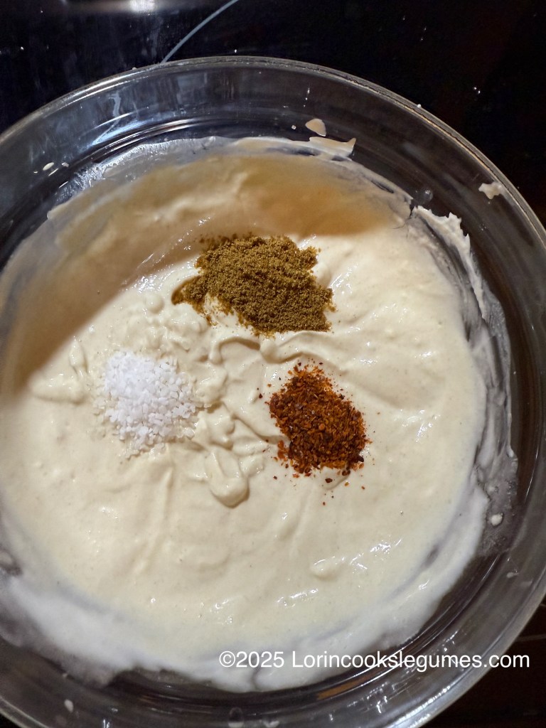 A bowl of tahini and yogurt sauce with piles of spices ready to be mixed in, including cumin, salt, and Aleppo pepper.