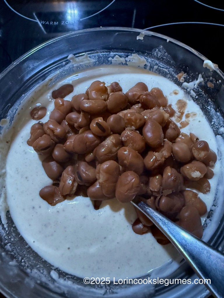 A bowl containing a creamy tahini and yogurt sauce, with a spoon resting inside and fava beans on top, ready to mix in. 