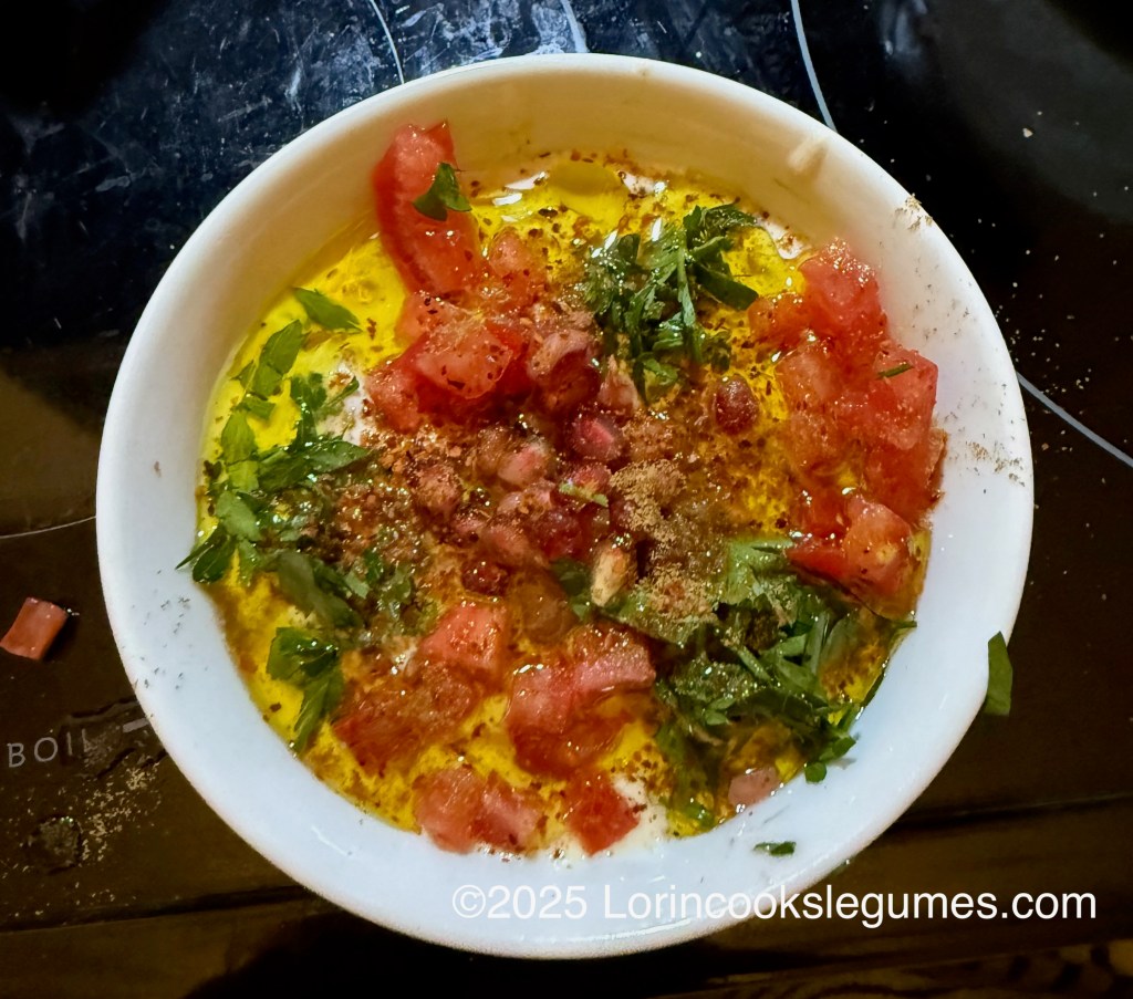 A bowl of Ful Medames topped with diced tomatoes, chopped parsley, pomegranate, ground cumin, and a generous drizzle of olive oil, set against a dark surface.