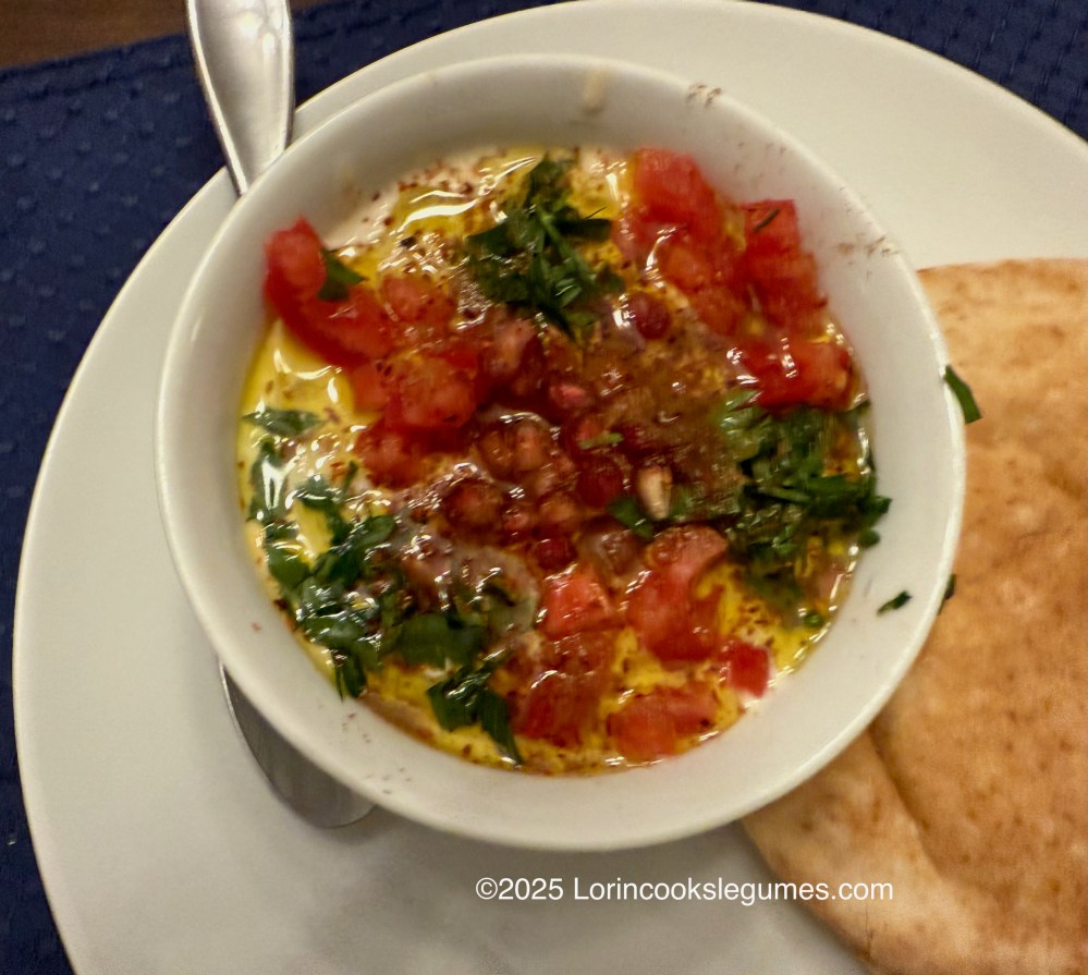 A bowl of Syrian ful medames topped with diced tomatoes, fresh parsley, pomegranate, and olive oil, accompanied by pita bread.