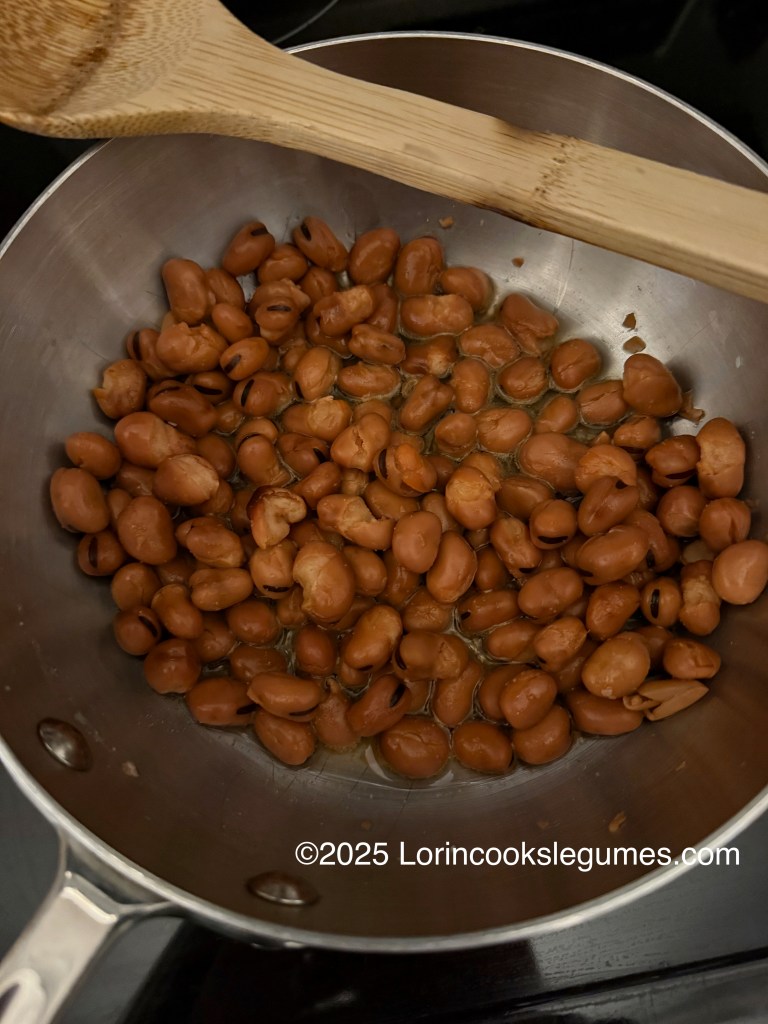 A pot containing cooked fava beans with a wooden spoon resting against the edge.