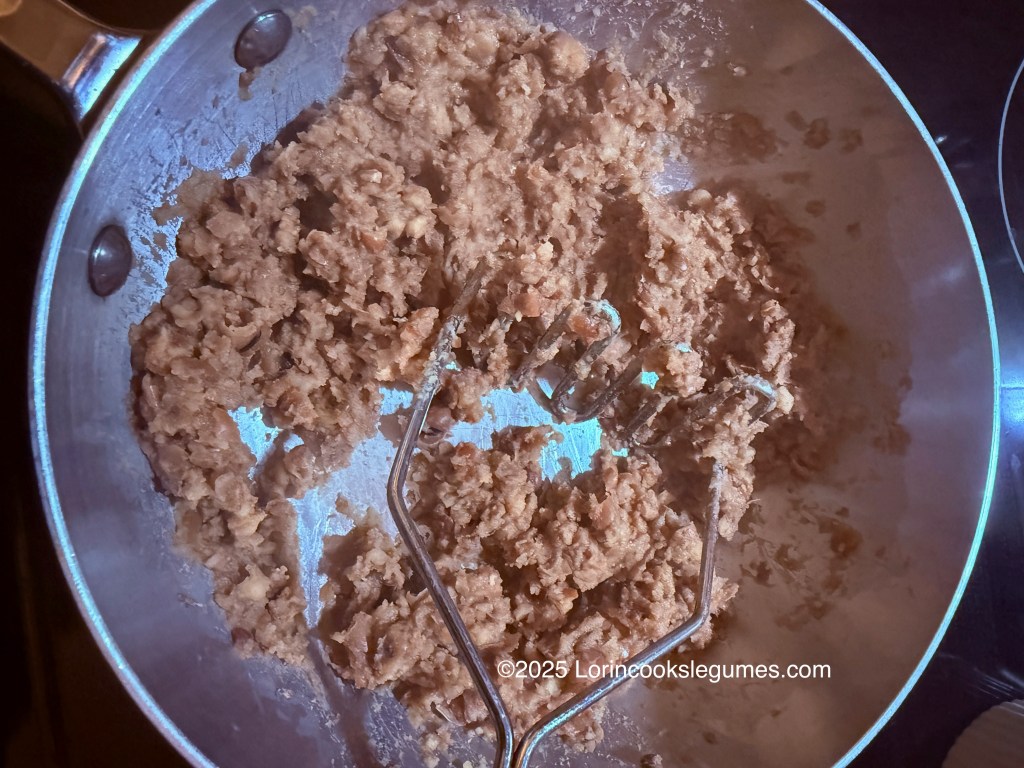 Mashed fava beans being prepared in a stainless steel pan with a masher.
