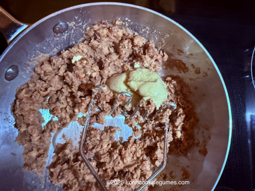 A close-up image showing a mixture of mashed fava beans in a pan with a spoonful of tahini added on top.