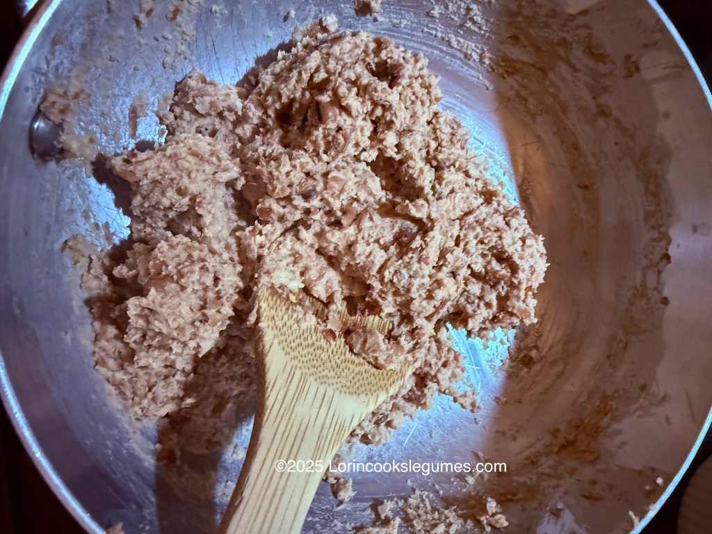 A bowl containing a thick, mashed mixture of fava beans, resembling hummus, with a wooden spoon partially embedded in it.