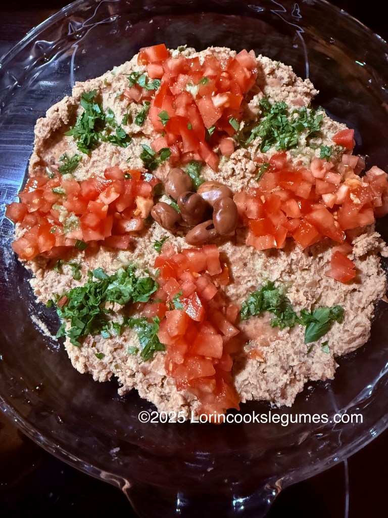 A glass plate of ful dip topped with diced tomatoes and fresh herbs, garnished with whole fava beans.
