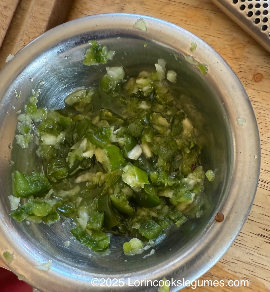 A metal bowl containing a mixture of minced green chilies and garlic, with some liquid at the bottom, placed on a wooden surface.