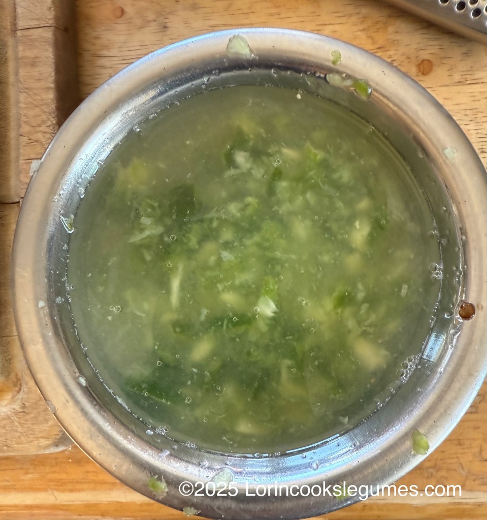 A metal bowl containing a mixture of garlic, green chilies, and lemon juice, showcasing a light green color with some bubbles on the surface, placed on a wooden surface.
