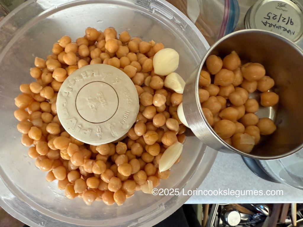 Drained chickpeas and garlic cloves in a food processor, arranged for hummus preparation.A small bowl of chickpeas reserved for garnish sits on the rim. 