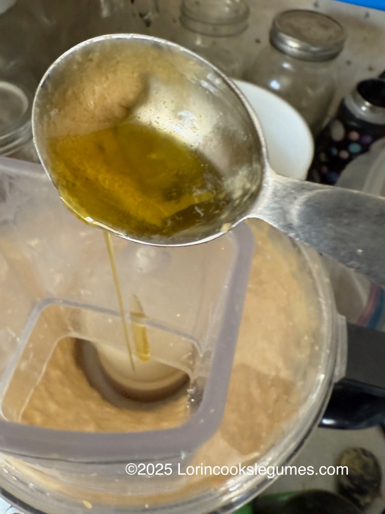 A metal tablespoon pouring olive oil into a blender filled with hummus, surrounded by jars and kitchen items in the background.