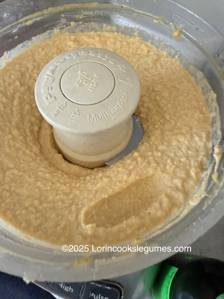 A close-up view of hummus in a food processor, showcasing a creamy and smooth texture. A mark in the surface shows that a finger has been used to scoop up a bit of hummus for tasting. 