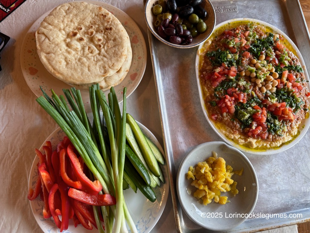 A spread featuring a dish of ful medames qudsiyah with layers of hummus and ful topped with diced tomatoes, chickpeas, and herbs, accompanied by pita bread, sliced vegetables including red peppers, cucumbers, and green onions, and  small bowls of olives and pickled cauliflower..