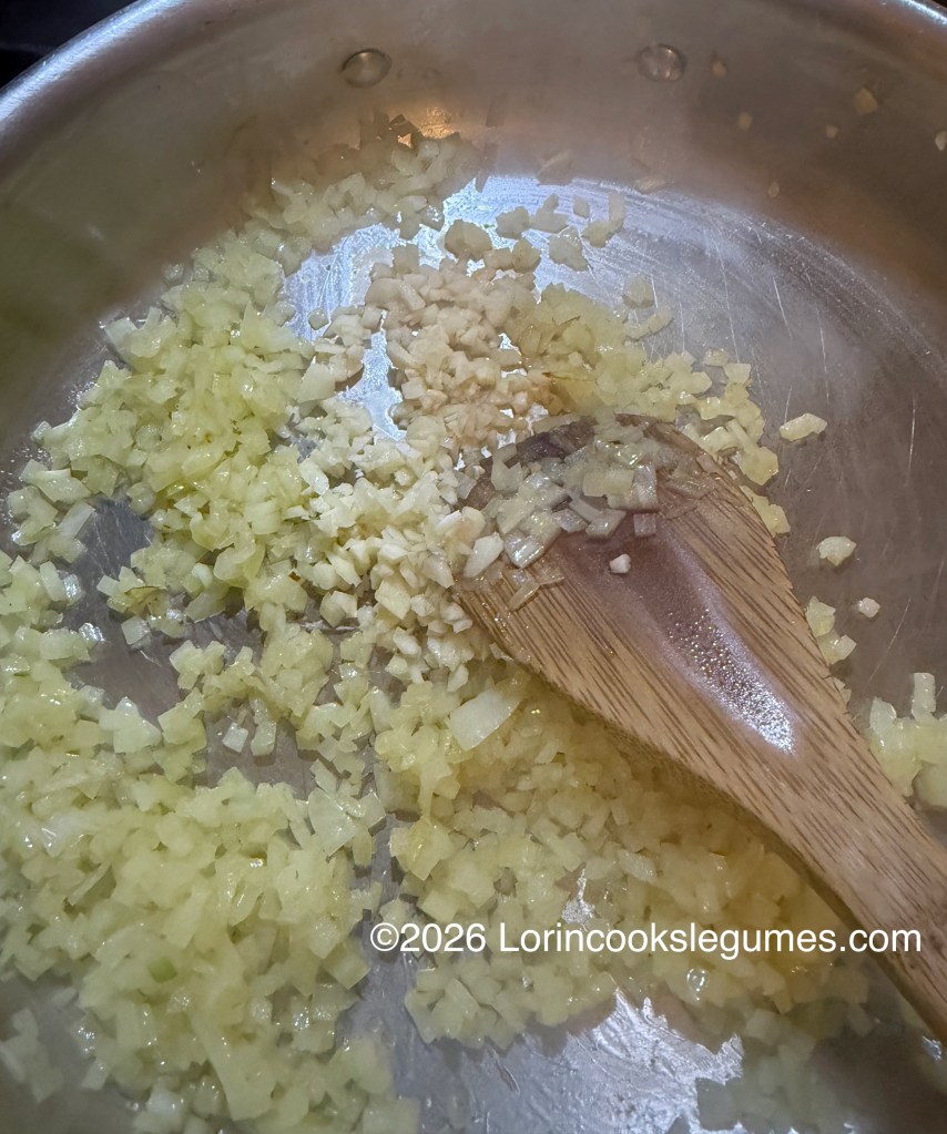 Chopped onions and garlic sautéing in a stainless steel pan with a wooden spoon.