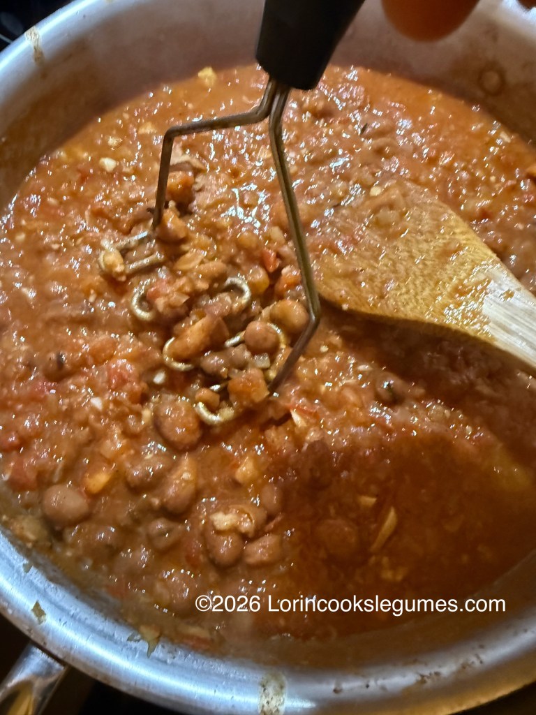 A close-up view of a pot filled with a chunky bean and tomato stew, with a potato masher standing in the pan and a wooden spoon resting on the edge.