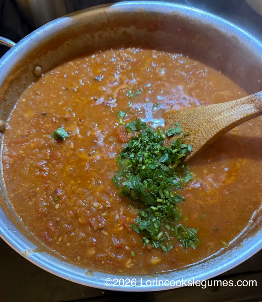 A pot of simmering soup with chopped cilantro being stirred in with a wooden spoon.
