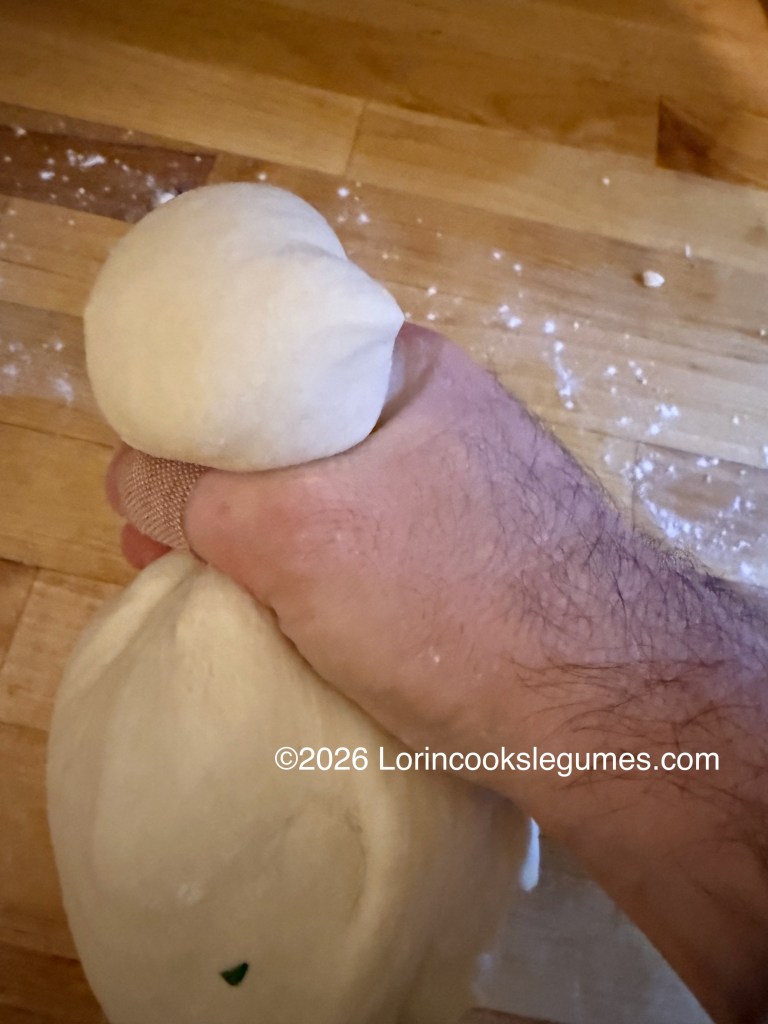 A hand holding a ball of dough, and pinching off a smaller ball, above a wooden countertop, with flour scattered around.