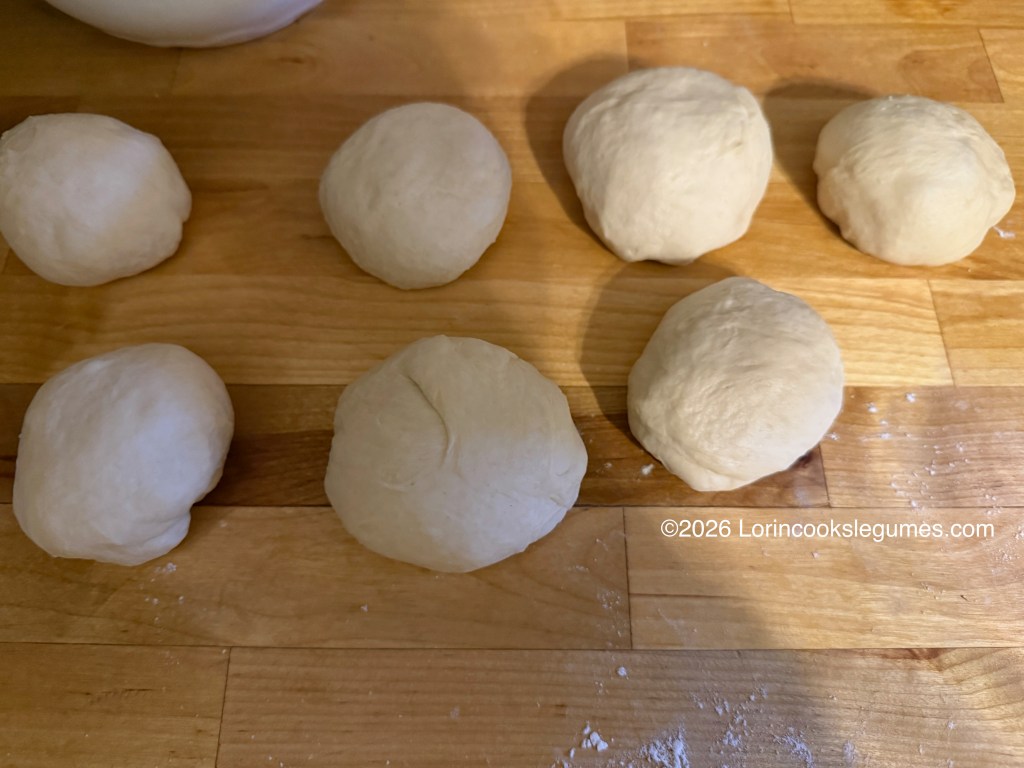 Seven round balls of dough resting on a wooden surface, with a light dusting of flour.