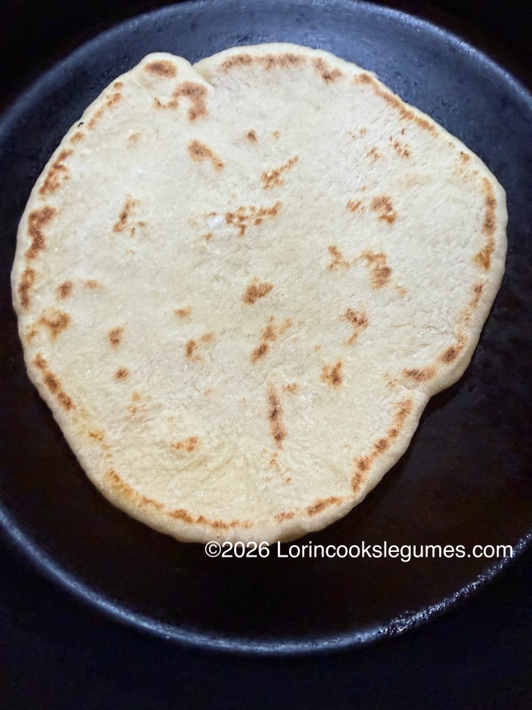 A golden-brown flatbread cooking in a skillet, showcasing a round shape with slight char marks.