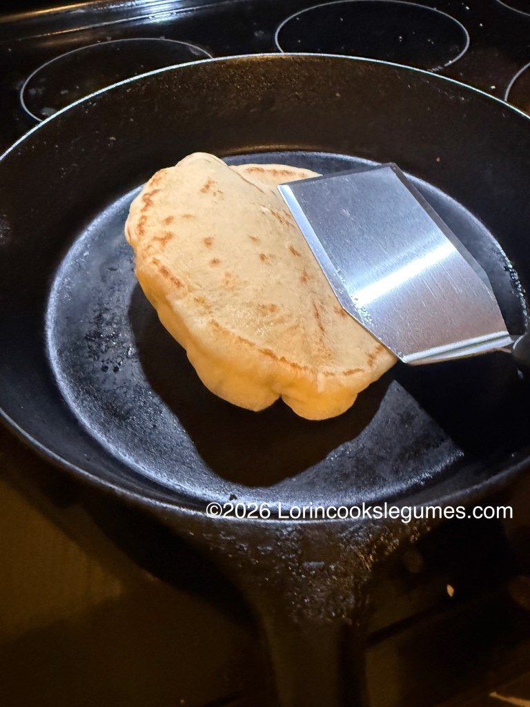 A skillet on a stovetop with a freshly cooked flatbread being pressed on one edge with a spatula, while the other edge puffs up. 