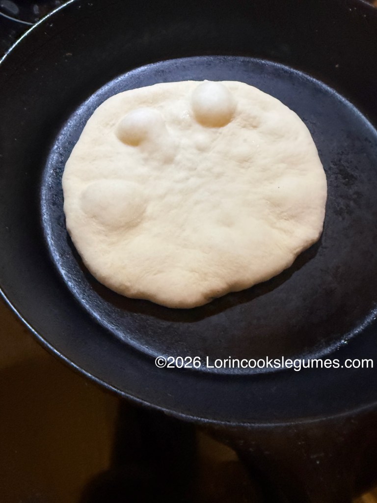 Raw dough disc in a hot frying pan, showing small air bubbles forming on the surface.