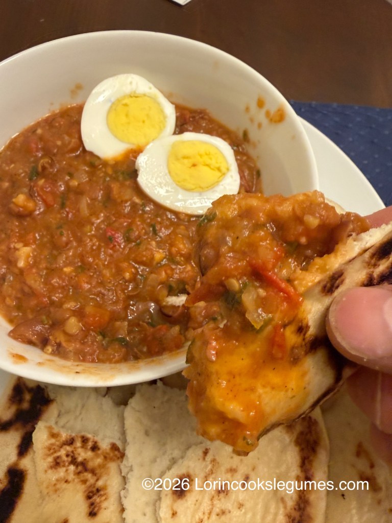 A hand holding a piece of flat bread that has been dipped in a bean stew, alongside a bowl of stew topped with a halved boiled egg.