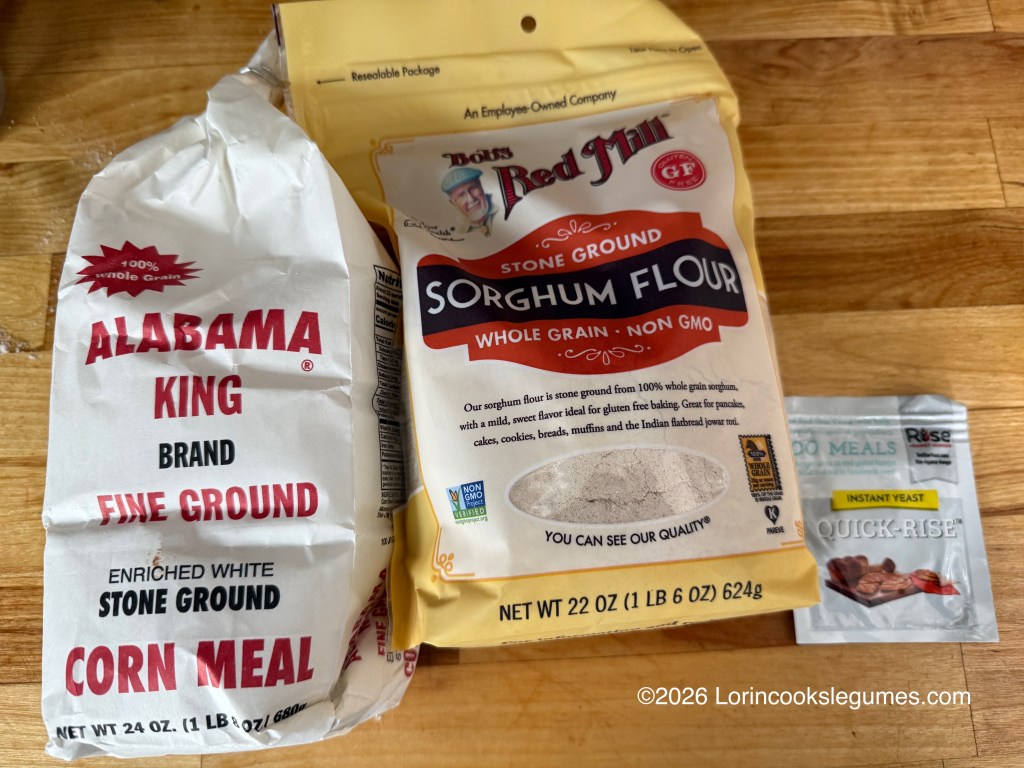 A package of Alabama King brand fine ground corn meal, a bag of Bob's Red Mill stone ground sorghum flour, and a sachet of quick-rise instant yeast on a wooden surface.