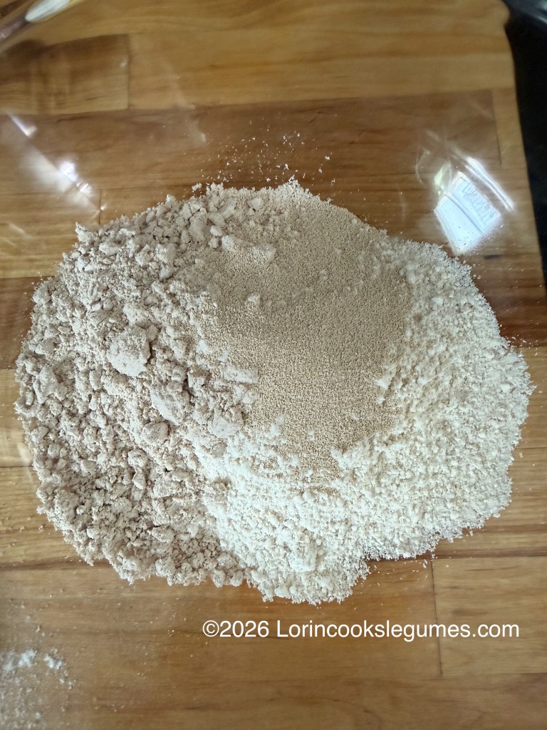 A pile of different types of flour and yeast in a glass bowl on a wooden surface.