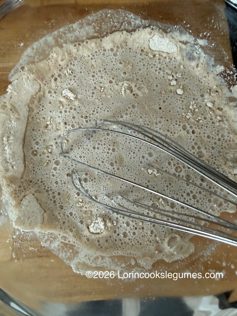 A glass bowl containing a bubbly mixture of water and flour, with a whisk resting inside.