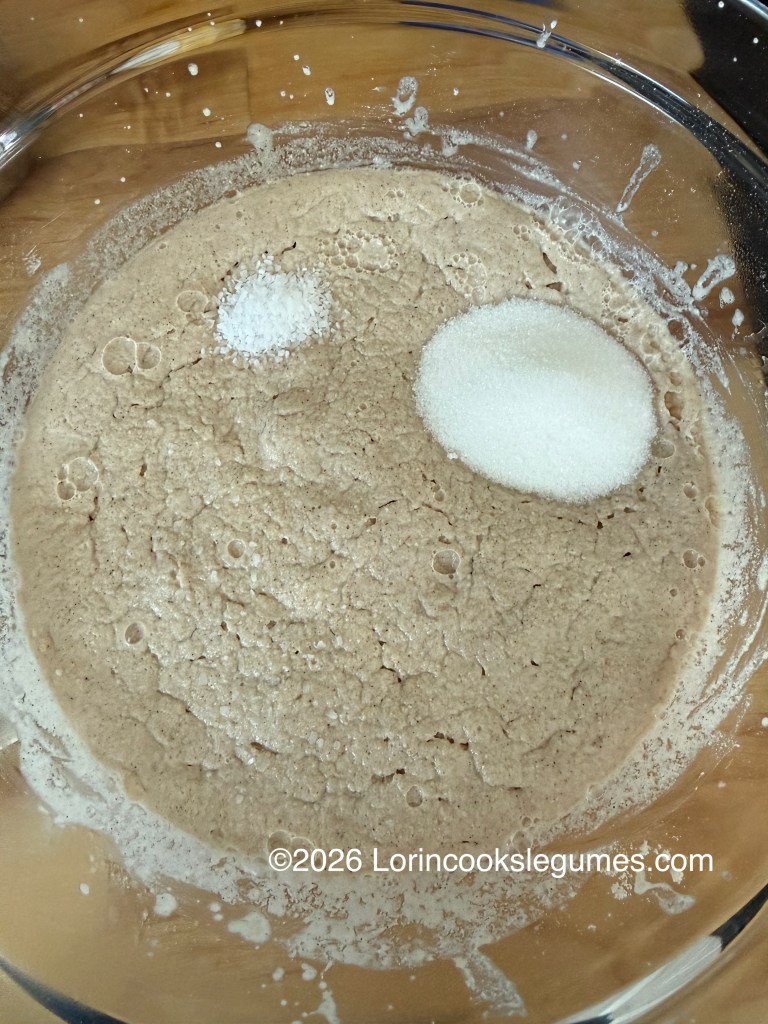 A clear glass bowl containing a bubbly mixture of dough, with two small piles of salt and sugar on top.