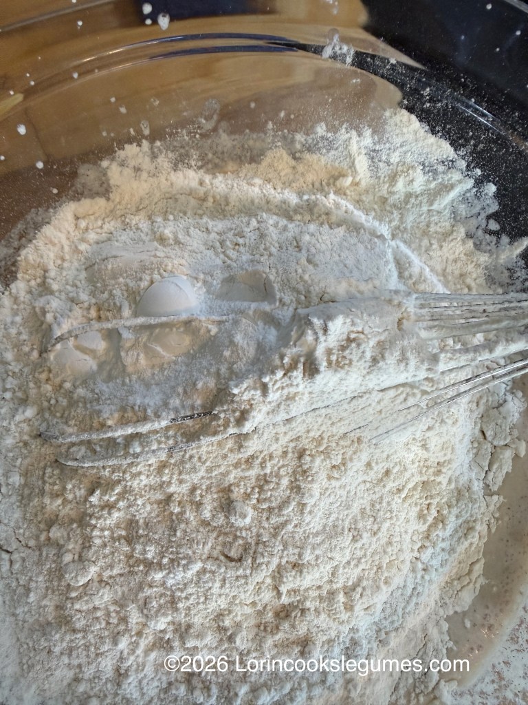 A glass bowl filled with flour and a whisk partially submerged in the mixture.