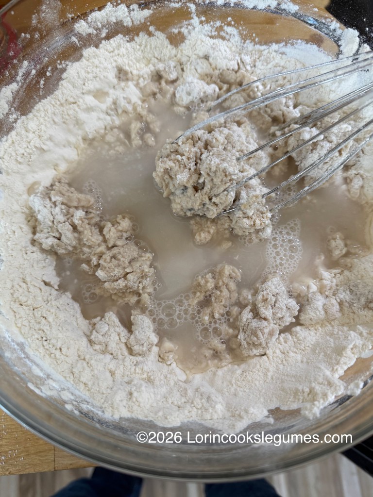 Close-up of a glass mixing bowl containing flour and water mixture, with clumps of dough and a whisk, placed on a wooden surface.