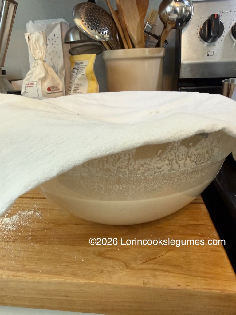 A bowl covered with a cloth, containing dough or batter, placed on a wooden countertop, surrounded by cooking utensils and ingredients.