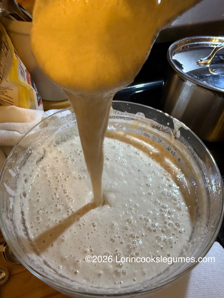 A ladle pouring thick, bubbly batter back into a large glass bowl filled with the same batter.